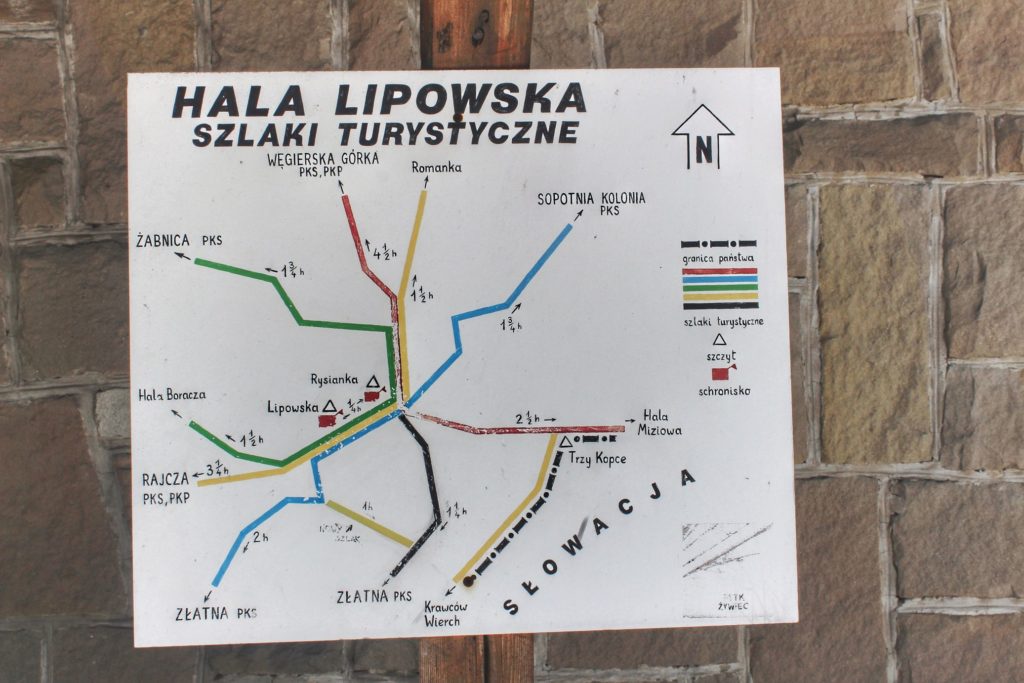 Biała tablica wisząca przy schronisku na Hali Lipowskiej opisująca przebieg szlaków turystycznych