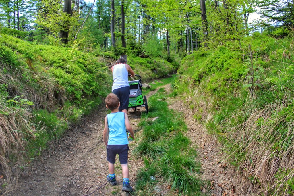 Letni dzień, leśna drog pnąca się w górę, dziecko oraz mężczyzna pchający wózek