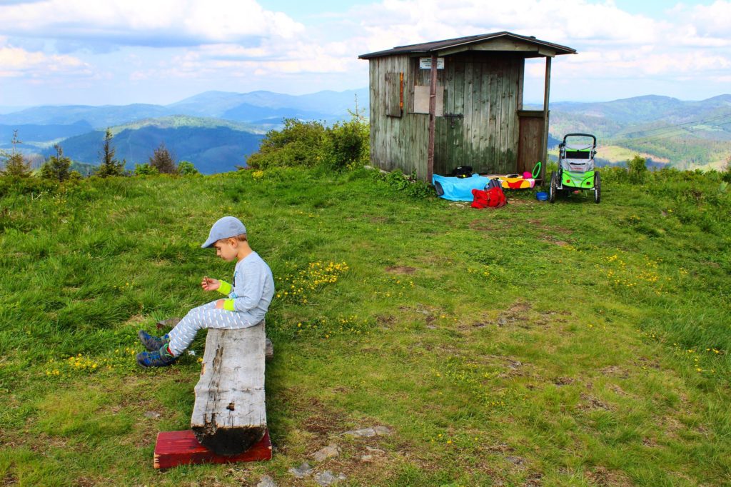 dziecko siedzące na drewnianej ławce na szczycie Wielka Racza, w tle drewniana buka nadleśnictwa obok której jest zielono szary wózek qeridoo na tle widoków górskich