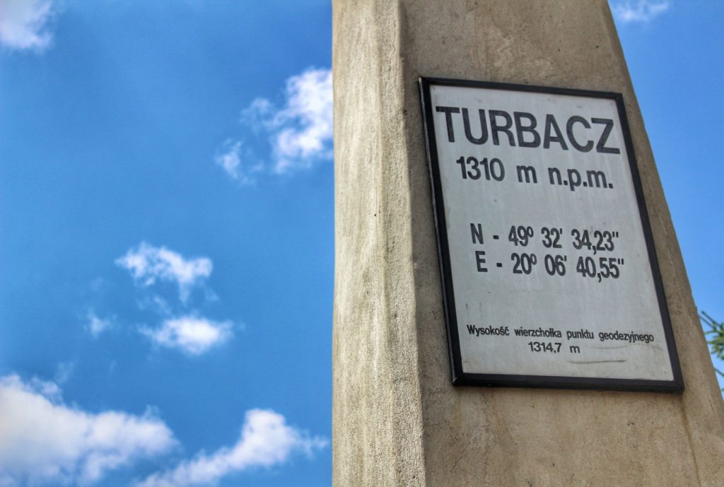 Biała tablica wisząca na betonowym słupie z napisem Turbacz 1310 m n.p.m.