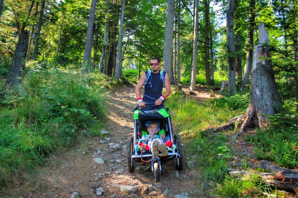Uśmiechnięty turysta z dzieckiem siedzącym w wózku, żółty szlak z Turbacza do Nowego Sącza, droga prowadząca przez las