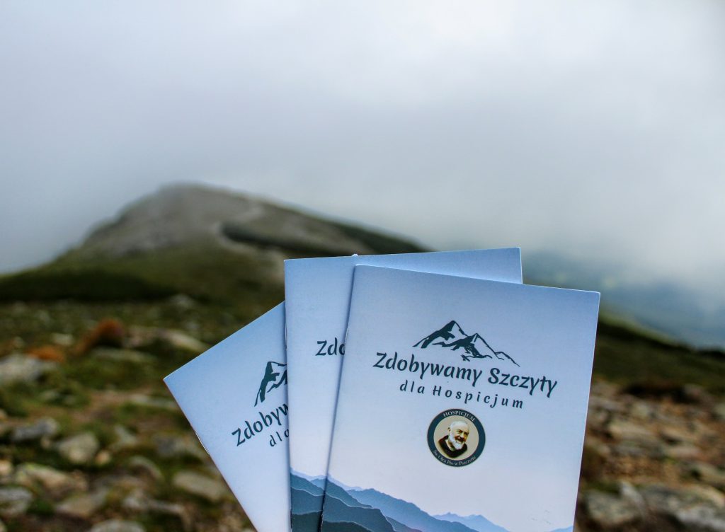 Trzy książeczki akcji Zdobywamy Szczyty dla Hospicjum na tle podejścia w paśmie Babiej Góry oraz zamglonego nieba