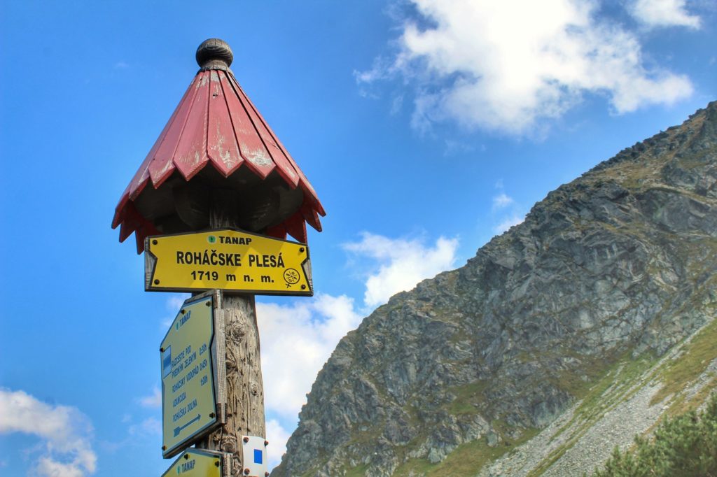 Tabliczka w kolorze żółtym z napisem Rohackie Stawy położone 1719 metrów nad poziomem morza, w tle tatrzańskie szczyty, niebieskie niebo