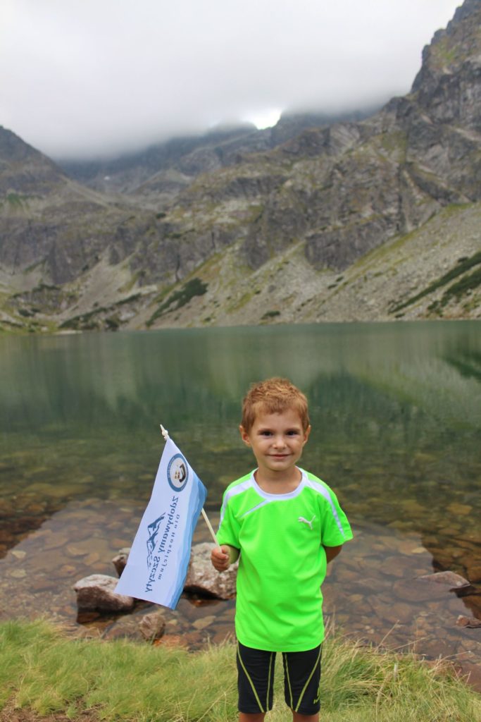 Zadowolone dziecko stojące na brzegu Czarnego Stawu Gąsienicowego w Tatrach Wysokich z flagą akcji Zdobywamy Szczyty dla Hospicjum