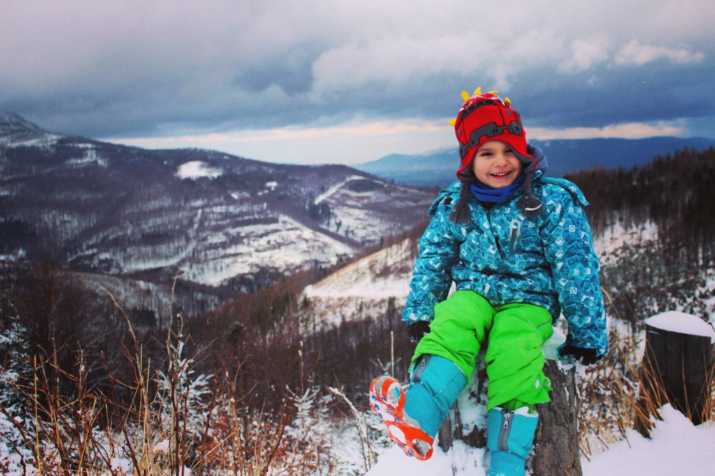 Uśmiechnięte dziecko siedzące na pniu drzewa, w koło zaśnieżone góry
