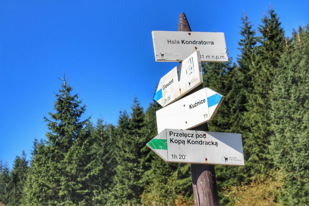 Biała tabliczka oznaczająca miejsce z napisem - Halla Kondratowa, drogowskazy szlak niebieski oraz zielony