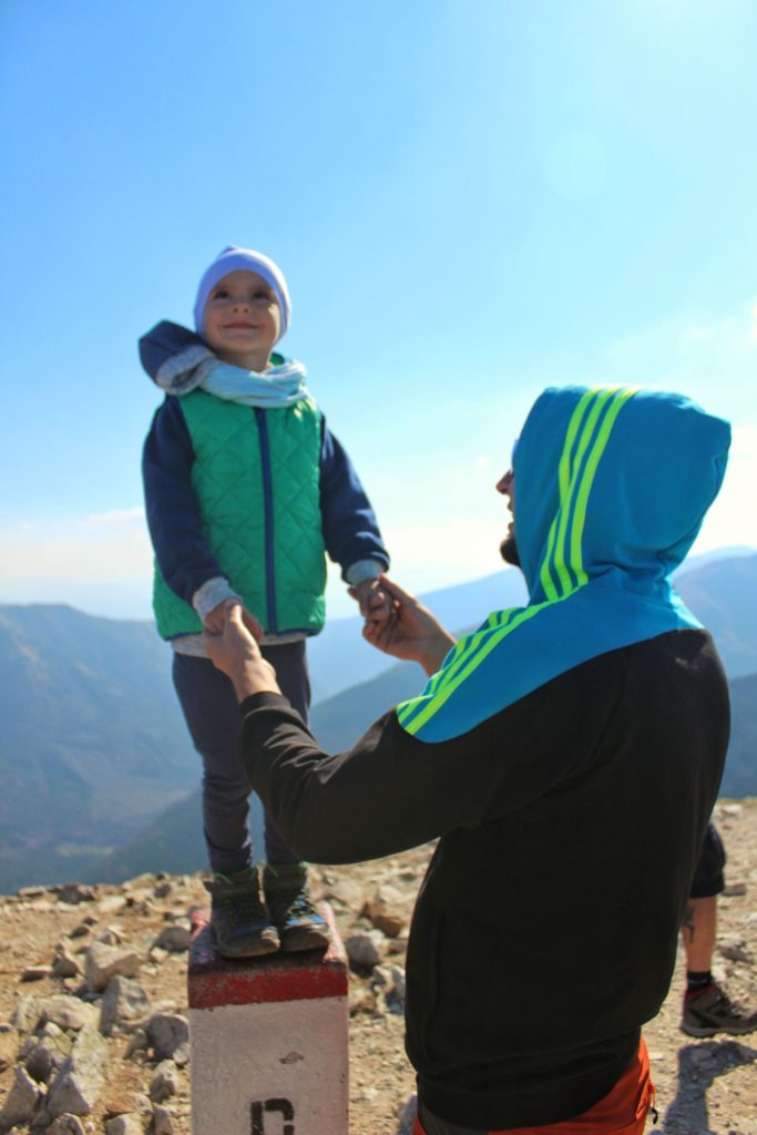 Uśmiechnięte dziecko patrzące w niebo, stojące na słupku granicznym na Kopie Kondrackiej, mężczyzna trzyma chłopca za rączki