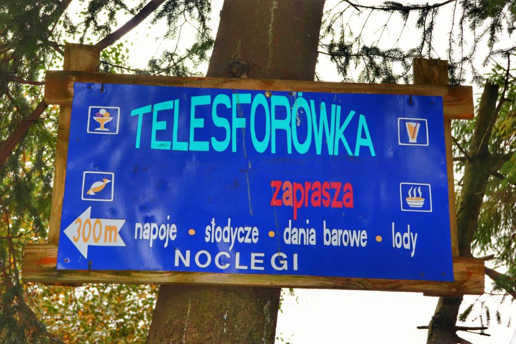 Niebieska tablica wisząca na drzewie na Trzech Kopcach Wiślańskich z napisem Telesforówka - 300 metrów, napoje, słodycze, dania barowe, lody, noclegi