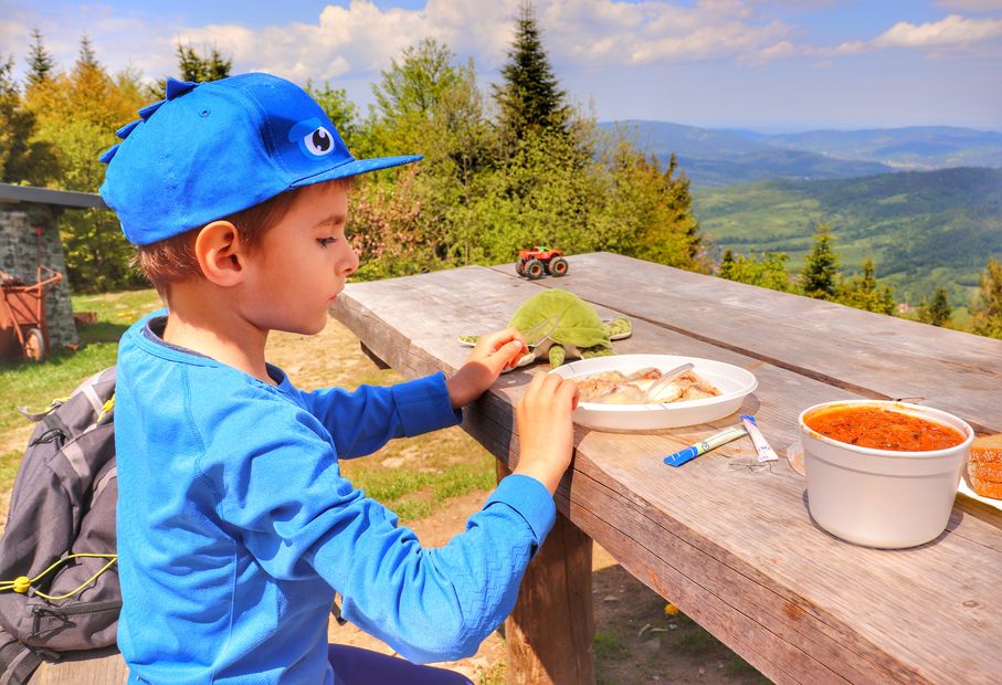 Dziecko siedzące na ławce przy drewnianym stole, jedzące pierogi zakupione w schronisku Luboń Wielki, w oddali krajobraz górski