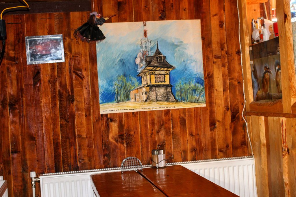 Wnętrze schroniska na Luboniu Wielkim, na drewnianej ścianie wisi obraz przedstawiający budynek schroniska