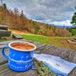 Niebieski, stalowy kubek turystyczny z białym napisem Karkonosze, trzy książeczki akcji Zdobywamy Szczyty dla Hospicjum, drewniany stół przy Bacówce na Wielkiej Rycerzowej