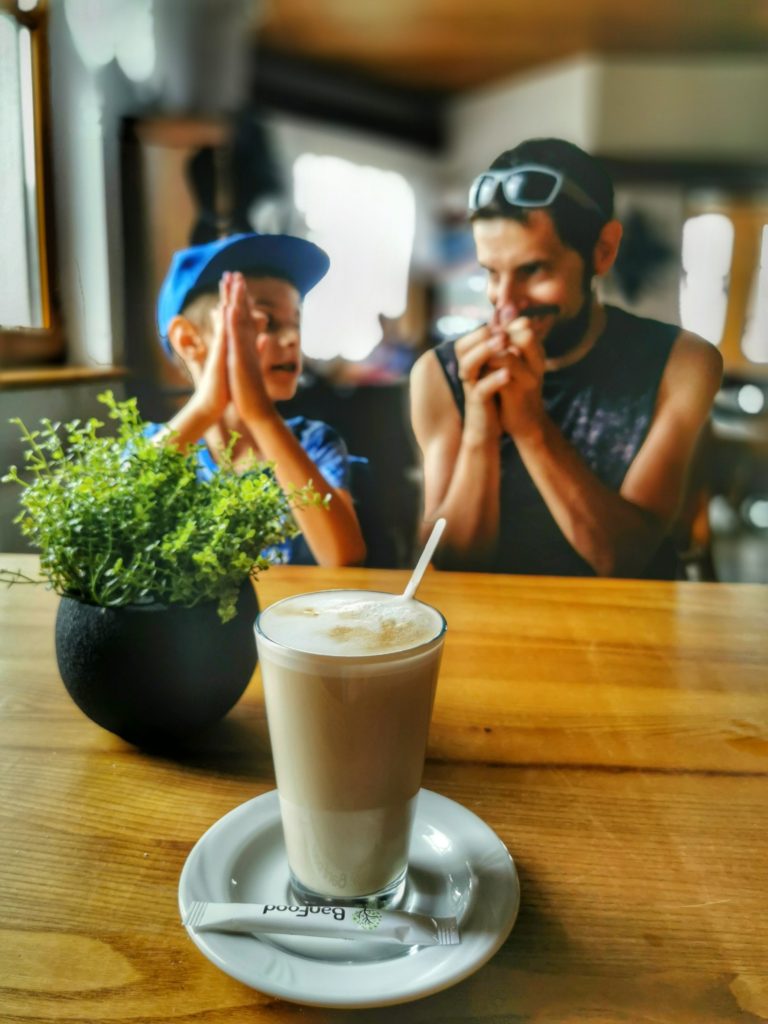 Szklanka, w której jest kawa latte, saszetka z cukrem na podstawku, w tle dziecko z tatą siedzący przy drewnianym stole w Schronisku na Magurce Wilkowickiej w Beskidzie Małym