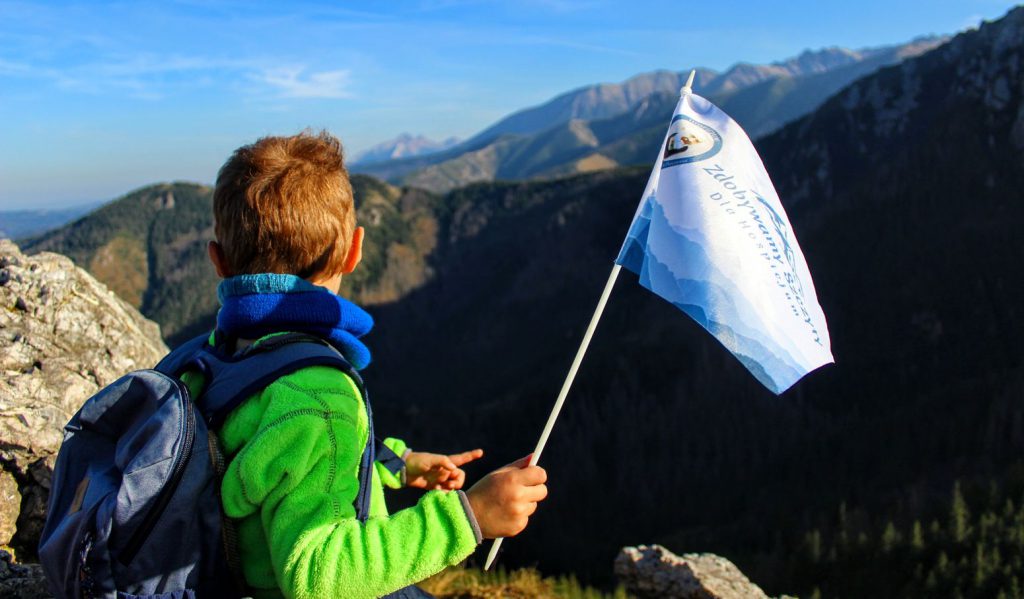 Dziecko na szczycie Sarnia Skała, obrócone tyłem, trzymające w rączce flagę akcji Zdobywamy Szczyty dla Hospicjum, w tle Tatry oraz błękitne niebo 