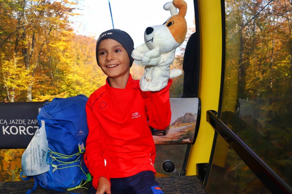 Uśmiechnięte dziecko w wagoniku kolejki linowej na Szyndzielnię trzymające maskotkę - pieska z bajki Reksia