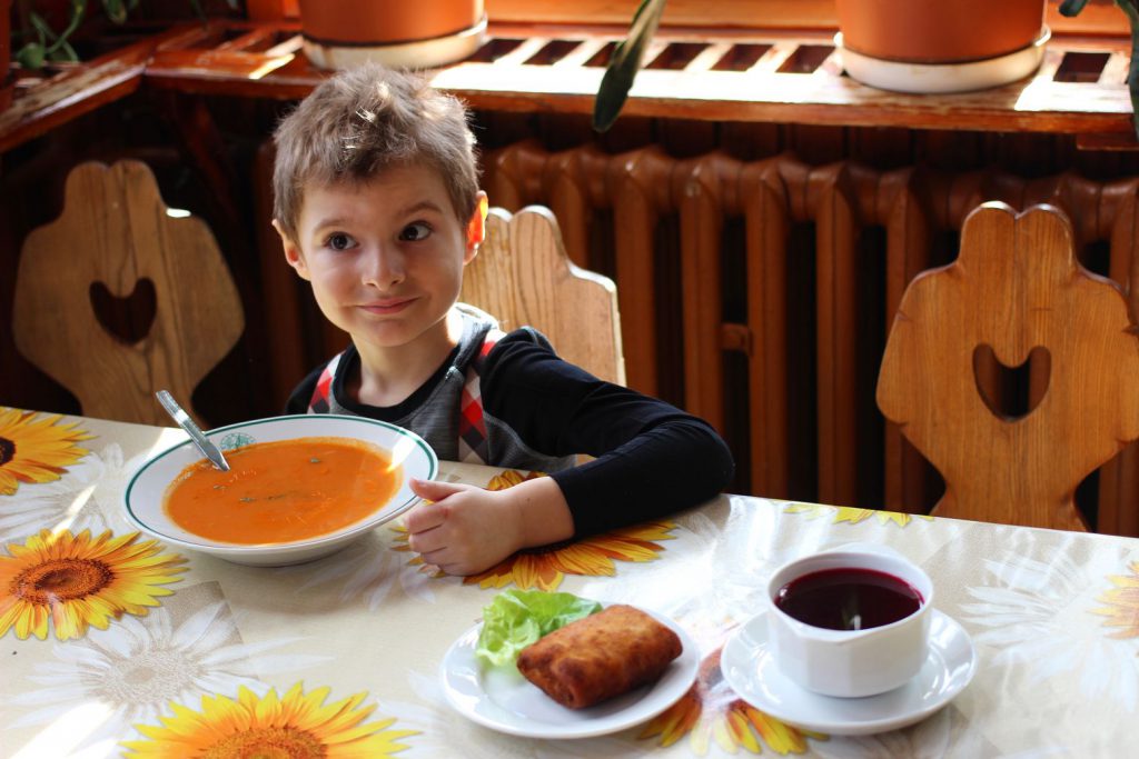 Uśmiechnięte dziecko siedzące w schronisku PTTK na Stożku przy stole z talerzem zupy pomidorowej oraz barszczem i krokietem 