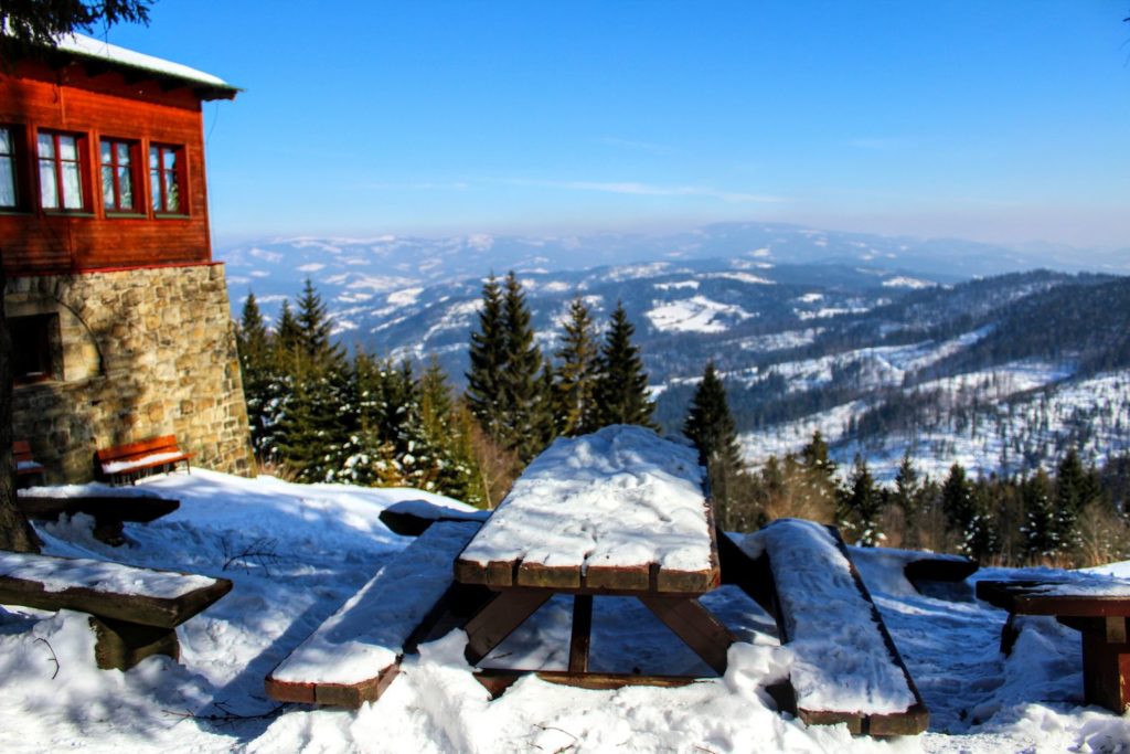 Widok na część budynku schroniska na Stożku Wielkim, stoły z ławkami, w tle krajobraz górski, zimowa sceneria 