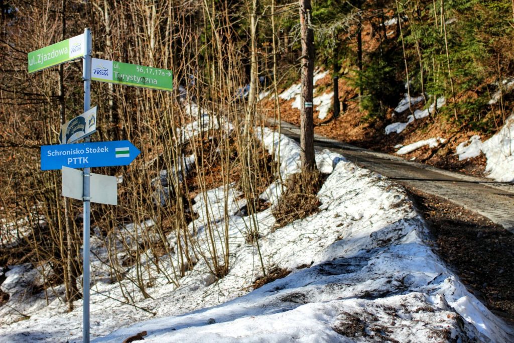 Słup z drogowskazami, niebieska tablica wskazująca drogę do Schroniska PTTK Stożek, śnieg zalegający na poboczu, droga