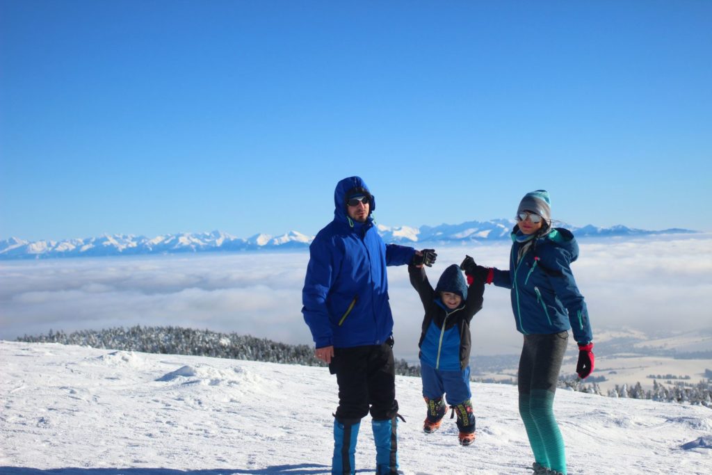 Trzyosobowa rodzina na szczycie Pilska zimą, matka z ojcem trzymają za ręce dziecko i podnoszą do góry, w tle tatrzańskie szczyty 