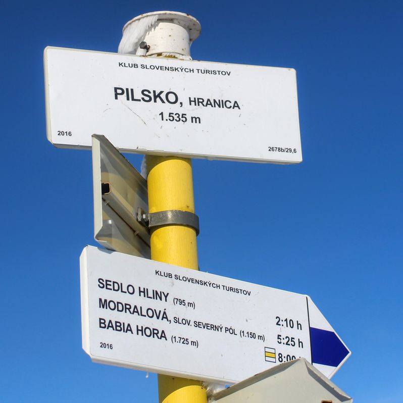 Biała tablica oznaczająca szczyt Pilsko wisząca na żółtym słupie, opis niebieskiego i żółtego szlaku na innej białej tabliczce