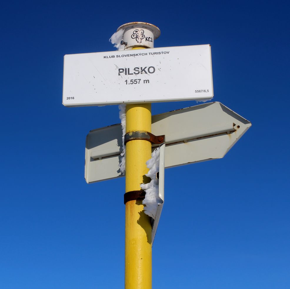 Biała tablica z napisem Pilsko 1557 metrów wisząca na żółtym słupie, niebieskie niebo 