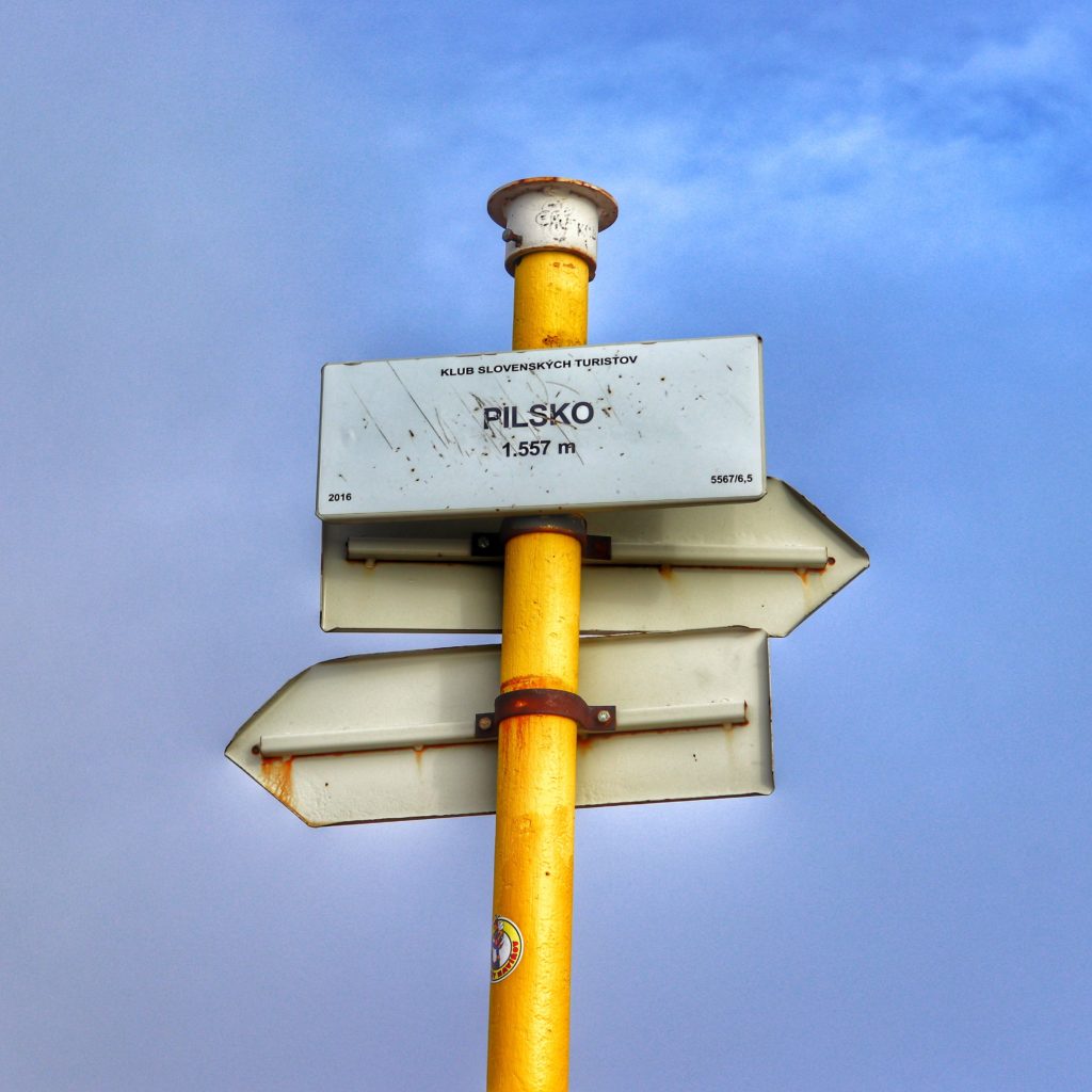 Biała tabliczka wisząca na żółtym słupie z napisem Pilsko 1557 metrów