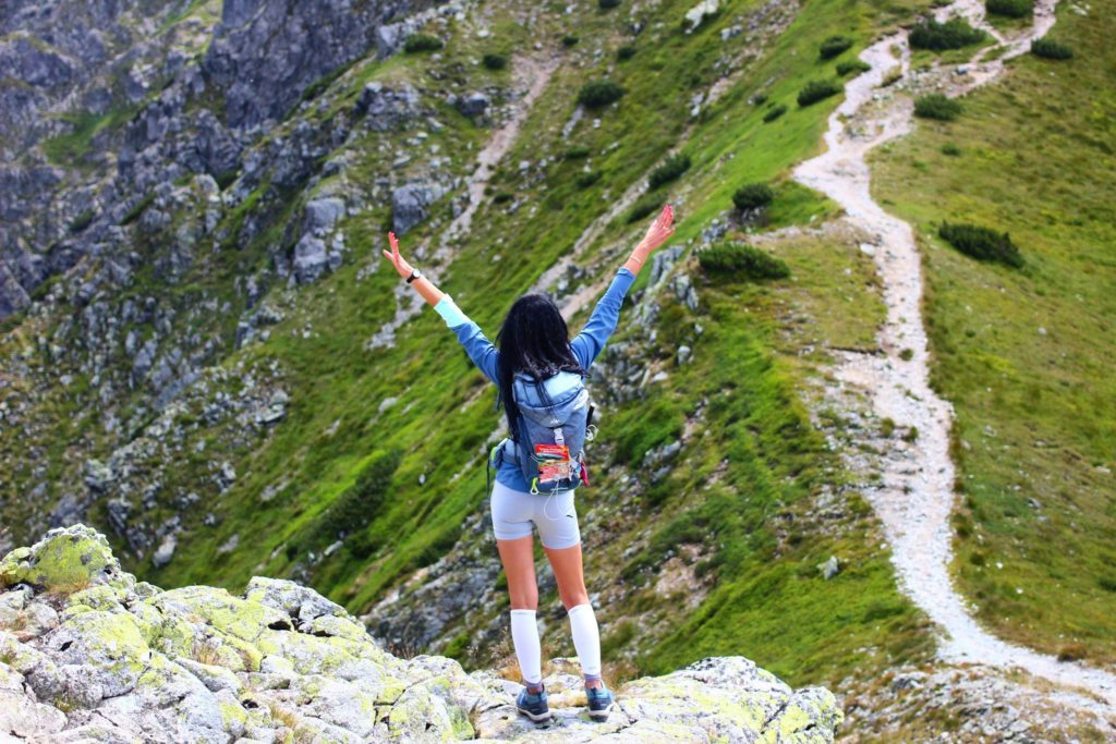 Kobieta obrócona tyłem z podniesionymi do góry rękami na Przełęczy Salatyńskiej, w koło skały oraz dróżka porośnięta zieloną trawą