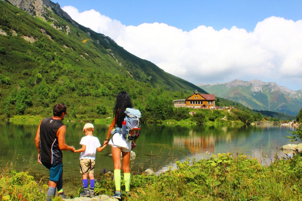 Trzyosobowa rodzina (po prawej mężczyzna, w środku dziecko i po lewej kobieta) stojąca tyłem na brzegu Zielonego Jeziora, w tle schronisko nad Zielonym Jeziorem odbijające się w tafli jeziora oraz okoliczne szczyty tatrzańskie