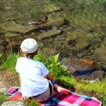 dziecko siedzące na kocu przy Zielonym Stawie Kieżmarskim patrzące na kaczkę krzyżówkę pływającą po stawie