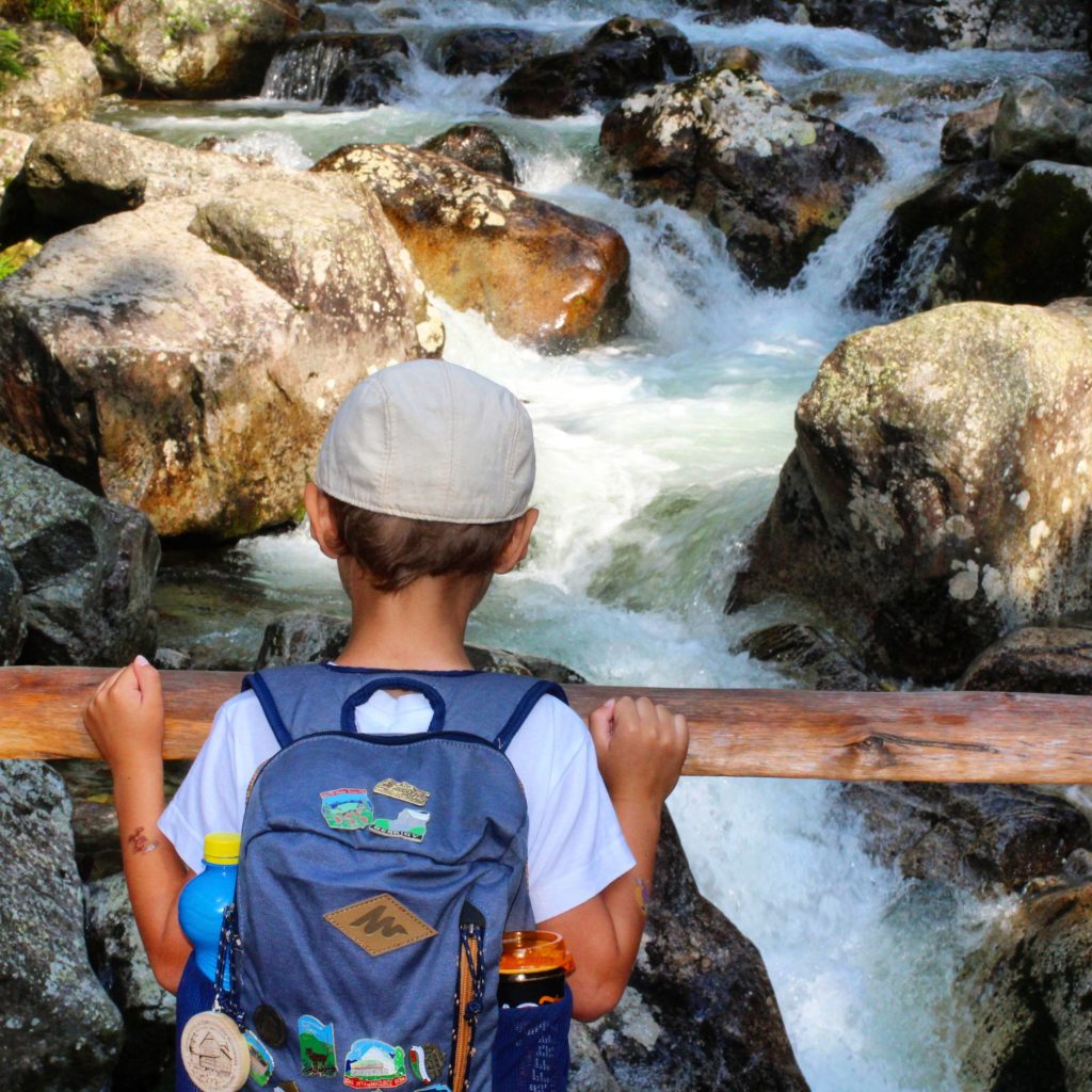 dziecko z plecakiem stojące tyłem na moście prowadzącym nad potokiem Biała Woda Kieżmarska, dziecko podziwia rwący potok oraz skały trzymając się drewnianej poręczy 