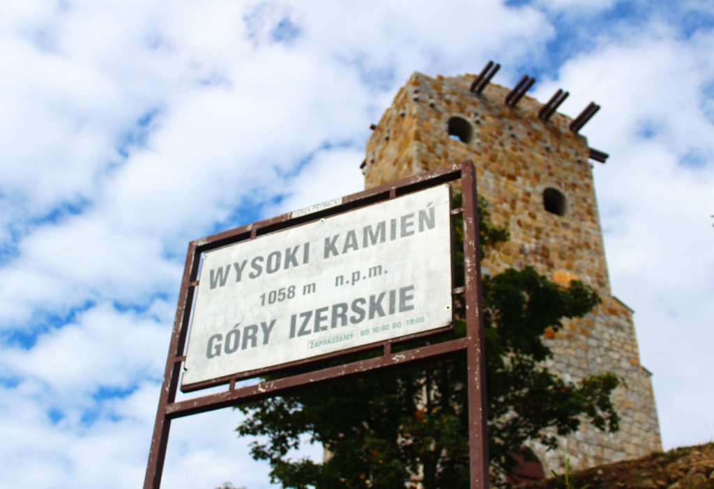 Biała tablica oznaczająca Wysoki Kamień leżący 1058 m n.p.m. w tle wieża widokowa na Wysokim Kamieniu