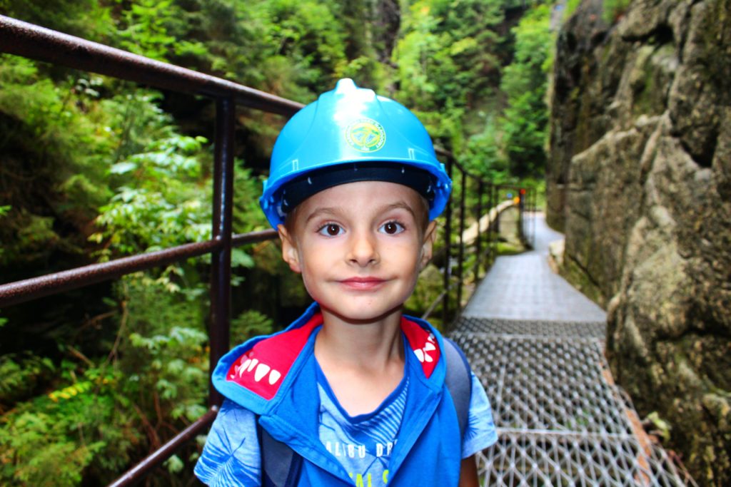 Dziecko w kasku Karkonoskiego Parku Narodowego stojący na metalowym moście obok skalnej ściany na trasie do Wodospadu Kamieńczyka