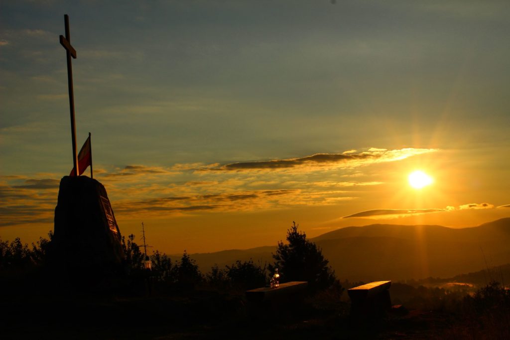 Pomnik, krzyż na szczycie Koczy Zamek oświetlony przez promienie wschodzącego słońca, w tle krajobrazy górskie