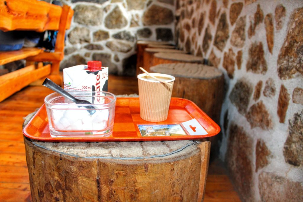 Wnętrze schroniska Wysoki Kamień, cukierniczka, mleko oraz kawa w kubku jednorazowego użytku leżąca na tacy, stole z pnia drzewa