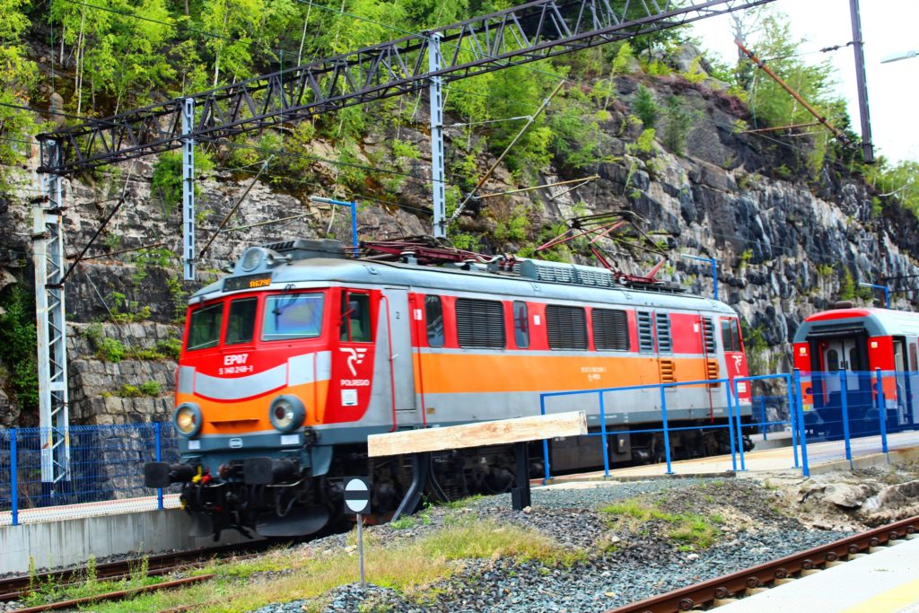 czerwono pomarańczowy pociąg stojący na stacji Szklarska Poręba Dolna, tory kolejowe oraz skalne ściany