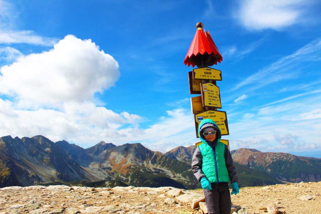 dziecko na tle tatrzańskich szczytów stojące pod słupem z żółtymi tabliczkami opisującymi szlaki żółty i niebieski oraz tabliczką oznaczająca szczyt Rakoń, niebieskie niebo z białymi obłokami