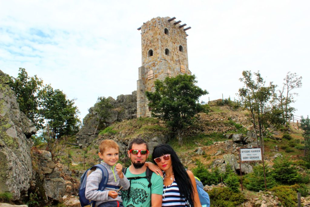 rodzina z dzieckiem na tle kamiennej wieży widokowej Wysoki Kamień oraz skał, biała tabliczka oznaczająca szczyt oraz informująca o terenie Gór Izerskich, 