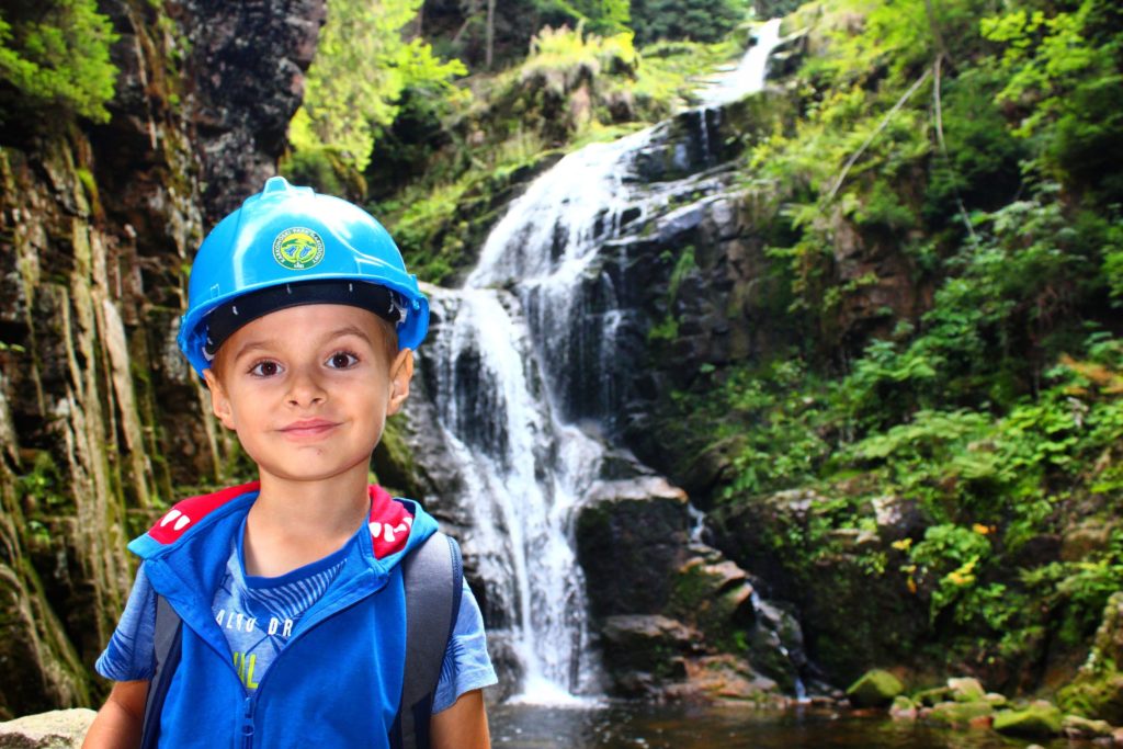 uśmiechnięte dziecko w niebieskim kasku Karkonoskiego Parku Narodowego na tle wodospadu Kamieńczyk, skał oraz zielonych drzew