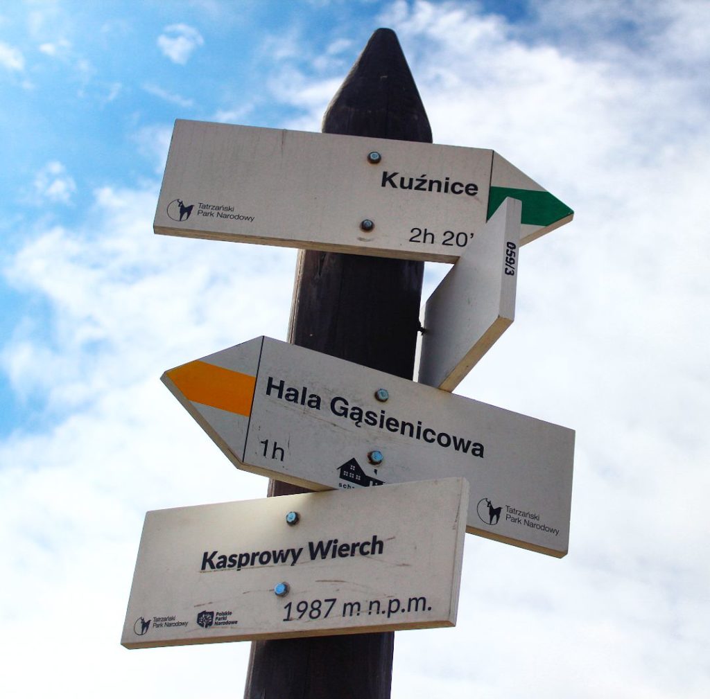 Słup z tablicą w kolorze białym oznaczającą Kasprowy Wierch mierzący 1987 m n.p.m. oraz drogowskazy opisujące szlak żółty na Halę Gąsienicową oraz szlak zielony do Kuźnic