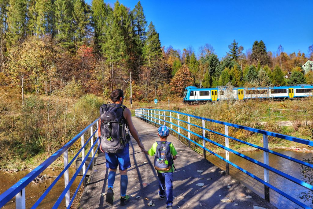 mężczyzna z dzieckiem przechodzący przez most nad rzeką Soła w Rajczy Dolnej, w tle widać nadjeżdżający pociąg, jesienna sceneria