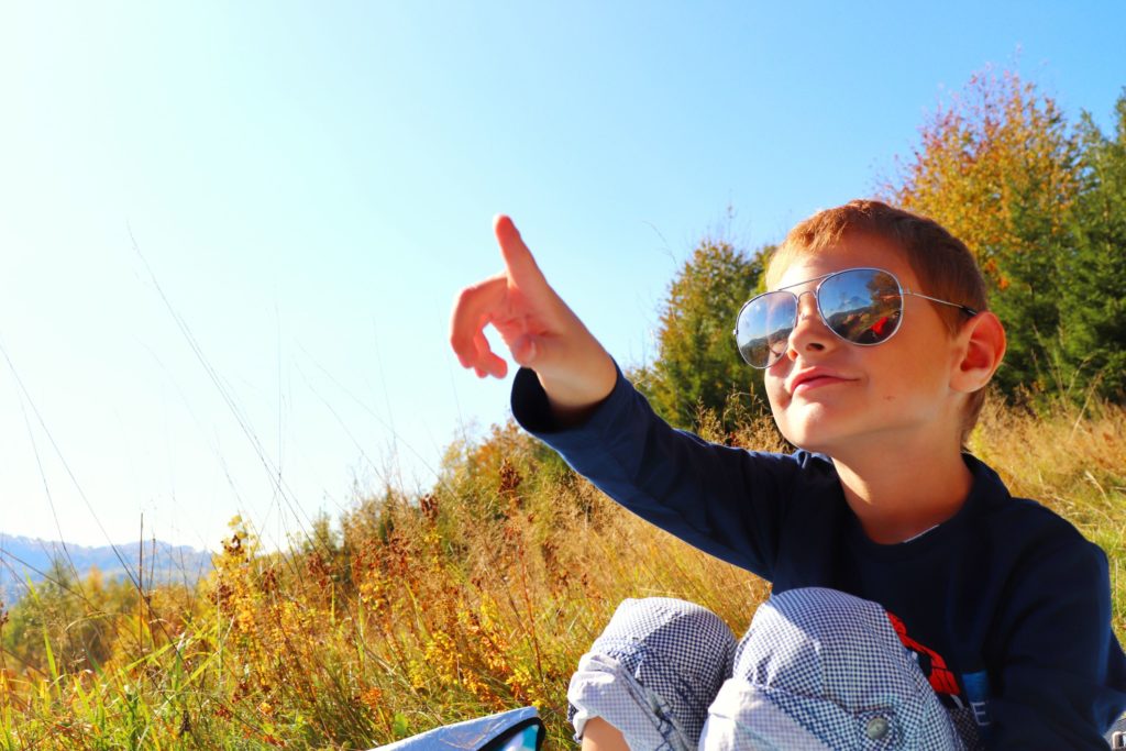 zadowolone dziecko w okularach słonecznych wskazujące palcem, siedzące na polanie, w koło jesień
