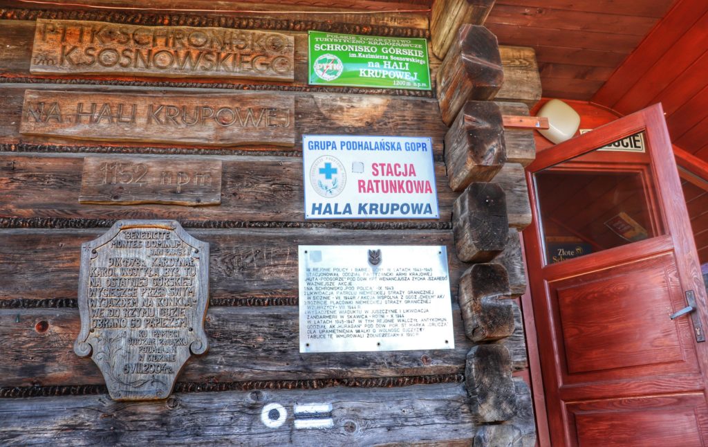 Drewniana ściana budynku Schroniska PTTK na Hali Krupowej, na której wiszą tablice, otwarte drzwi