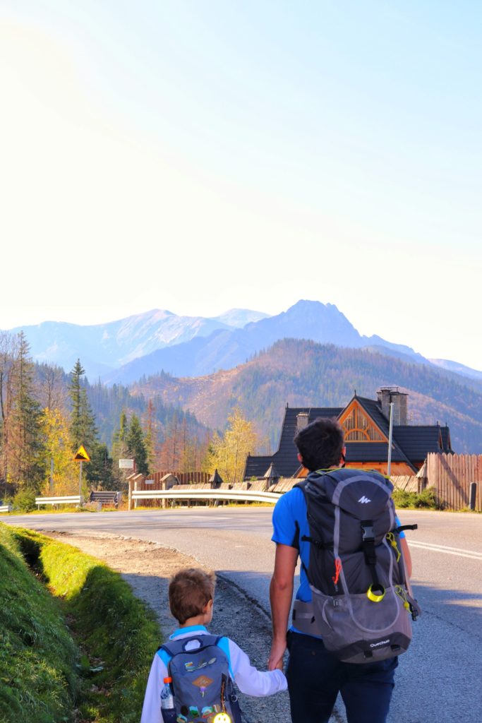 Dziecko idące za rękę z tatą lewą stroną ulicy - poboczem, w tle tatrzańskie szczyty