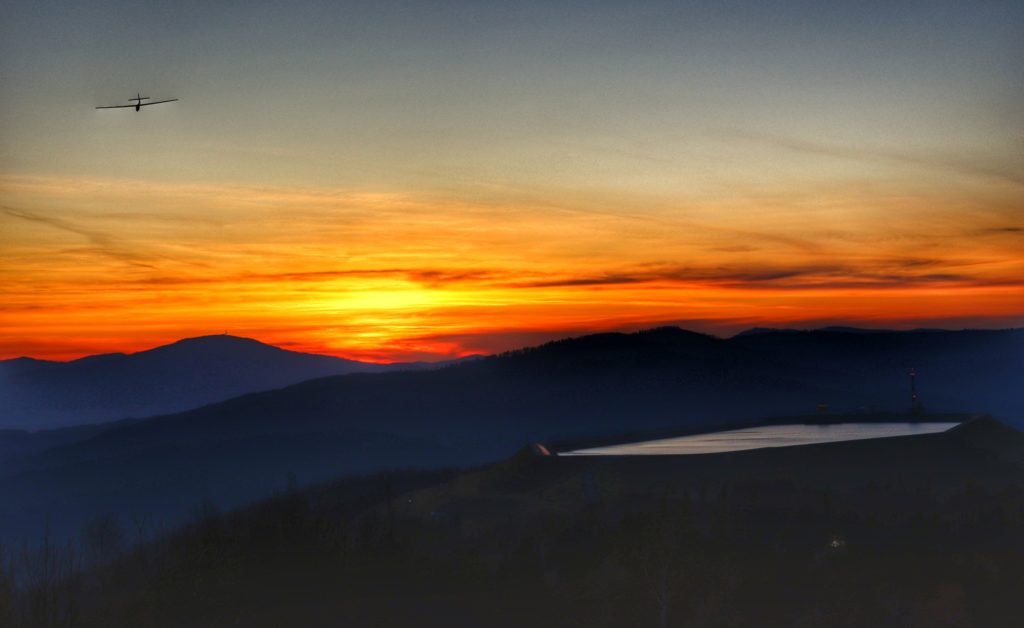 Szybowiec na niebie zakolorowanym przez zachód słońca, w tle widoki górskie, w tym Skrzyczne, po prawej zbiornik na Górze Żar