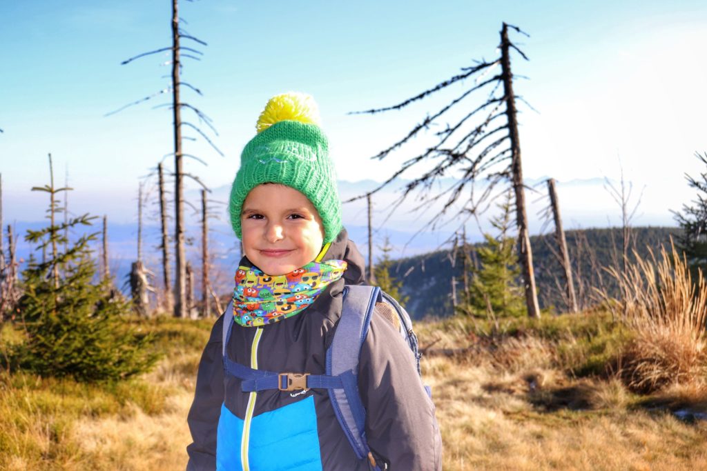 Zadowolone dziecko na szczycie Polica, w tle drzewa, w oddali, za mgłą Tatry