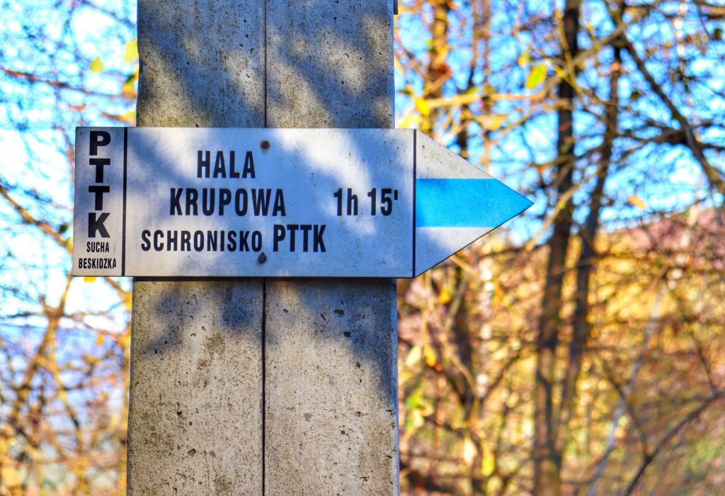 Biała tablica w kształcie strzałki opisująca niebieski szlak na Halę Krupową, czas przejścia według drogowskazu 1 godzina 15 minut