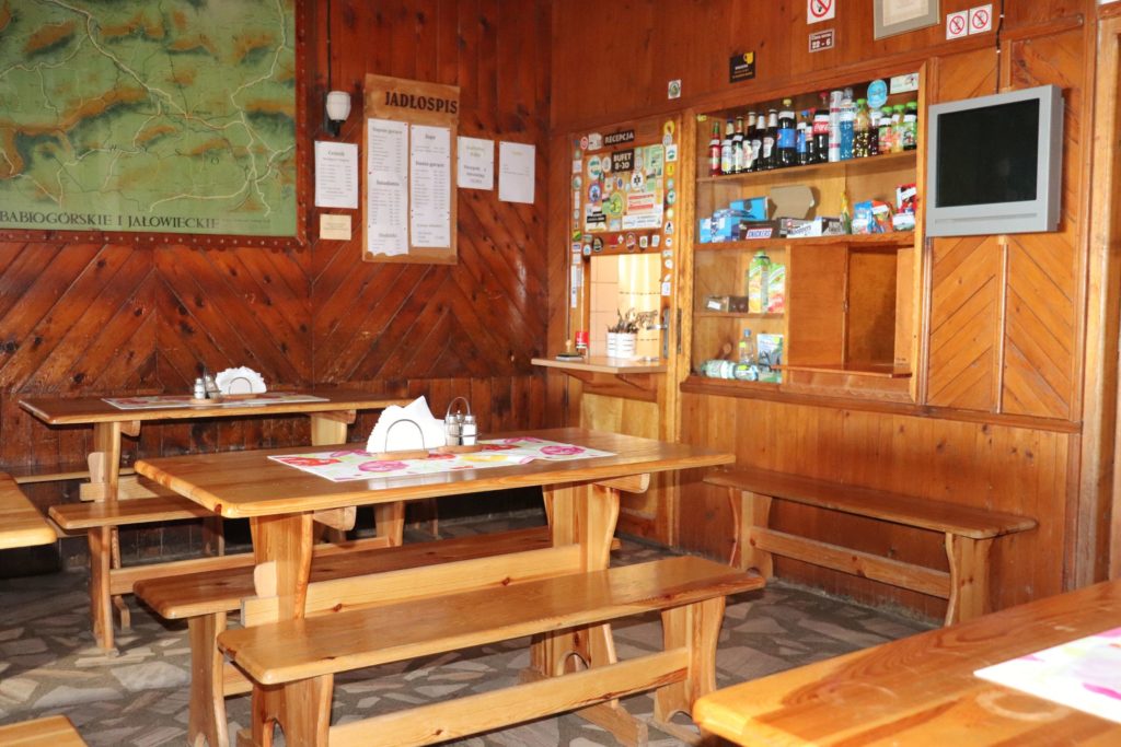 wnętrze schroniska na Hali Krupowej, drewniane stoły ławki, na śianie telewizor, bufet z wiszącym na ścianie jadłospisem