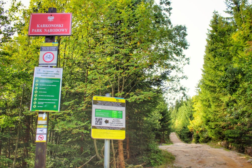 Czerwona tablica informująca o terenie Karkonoskiego Parku Narodowego, tablica z cennikiem Parku, obok szeroka droga