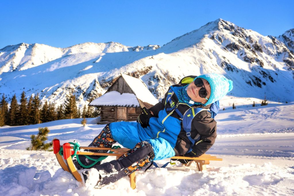 Dziecko na Hali Gąsienicowej zimą, zadowolony chłopiec siedzi na sankach na tle Tatr