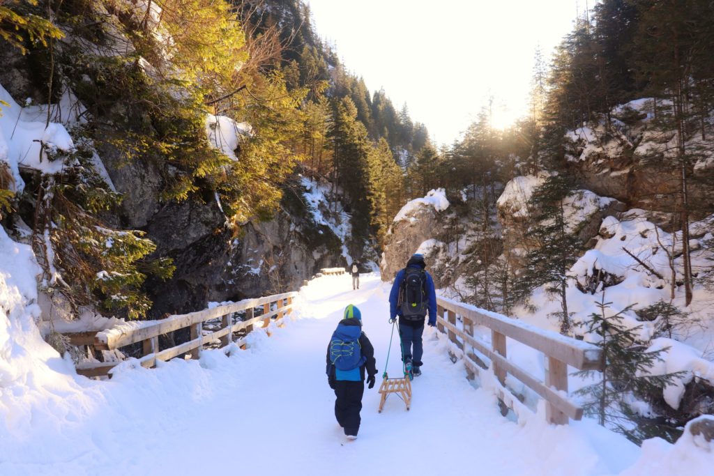 Turyści, w tym dziecko idące po zaśnieżonej drodze przez Wąwóz w Dolinie kościeliskiej, drewniany most, skalna ściana