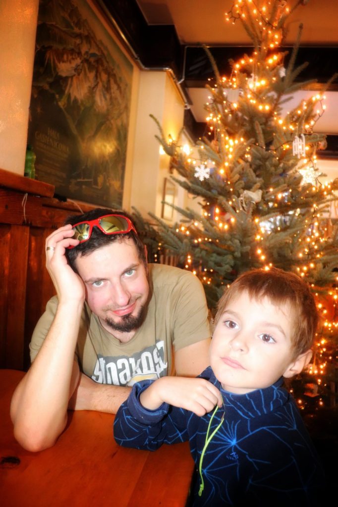 Uśmiechnięty mężczyzna, dziecko siedzący przy stole w Schronisku Murowaniec, z tyłu ubrane świąteczne drzewko